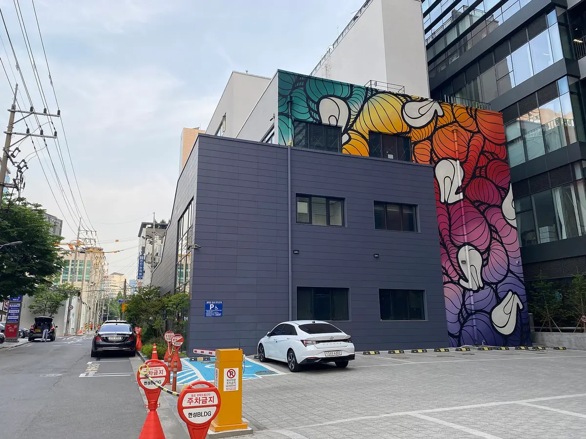 ציור קיר צבעוני על מבנה תעשייתי אפור בפינת רחוב, ומכוניות חונות לפניו